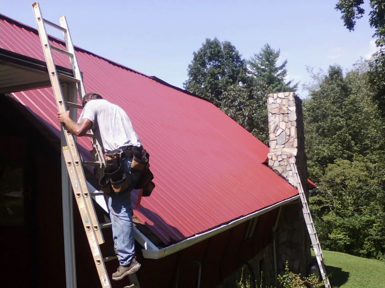 Roofer on a ladder beside a red metal roof and stone chimney — Soffit & Fascia Repair by Roofing Brothers in Madras